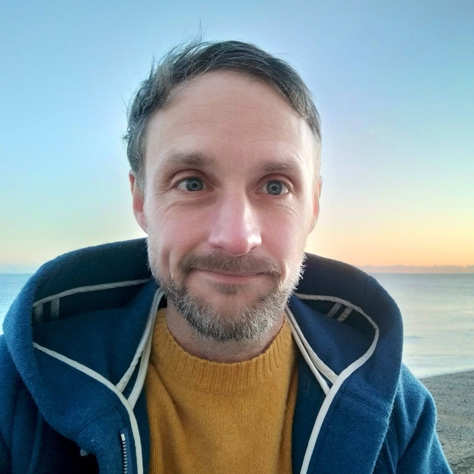 Rounded photo portrait of Sam Hawkins. Wearing a blue duffel and yellow jumper with the sea and a yellow and blue sunrise sky behind